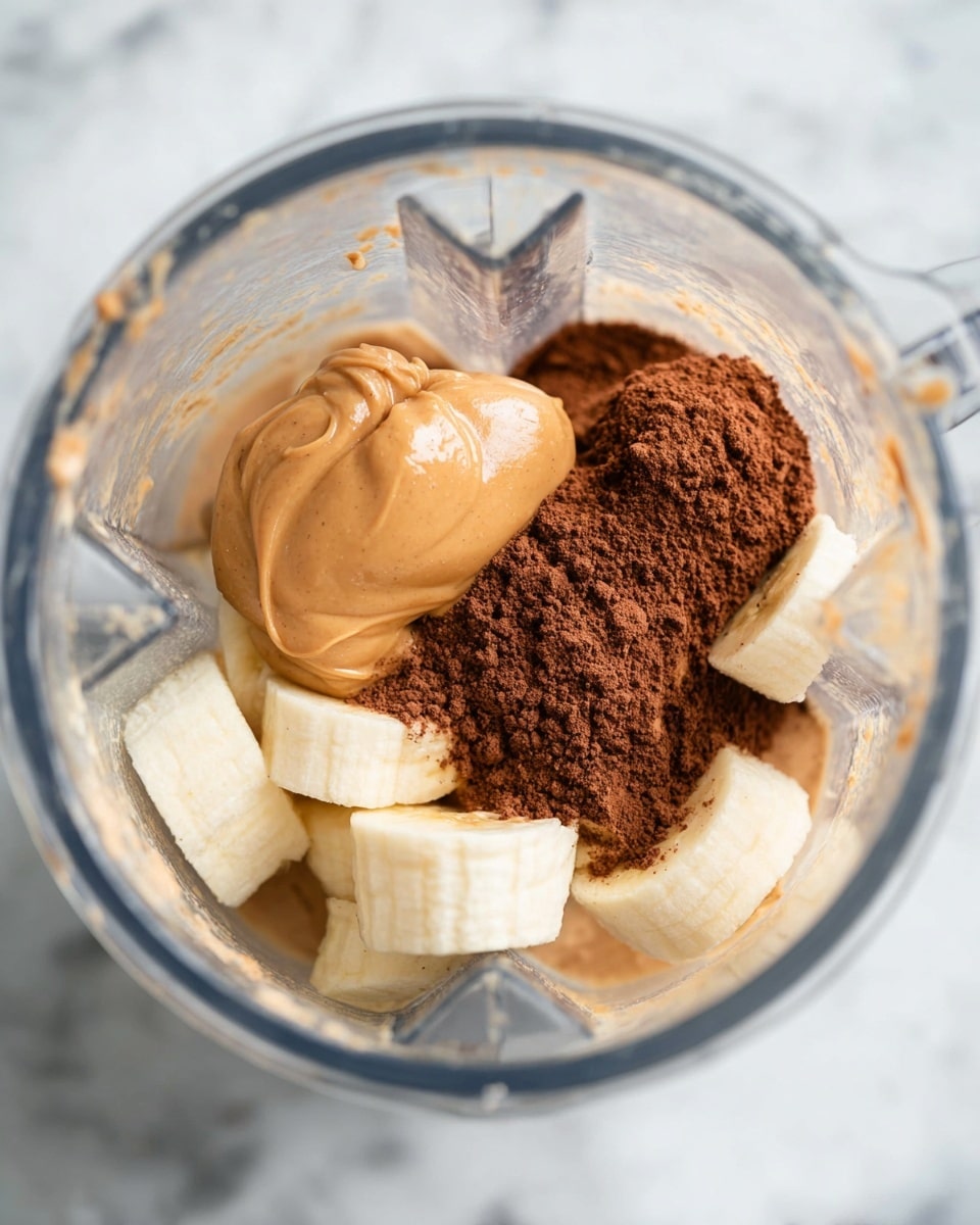 Inside a clear blender jar placed on a white marbled surface, there are several visible layers of ingredients. The bottom layer is a light beige or creamy liquid. On top of it, there are thick slices of pale yellow frozen banana pieces scattered around. Next to these banana pieces is a pile of fine, dark brown cocoa powder. Sitting above the banana and cocoa powder is a smooth dollop of peanut butter with a warm tan color and slightly shiny texture, resting in the center. The whole scene is captured in close-up, showing the texture and color of each ingredient clearly. photo taken with an iphone --ar 4:5 --v 7