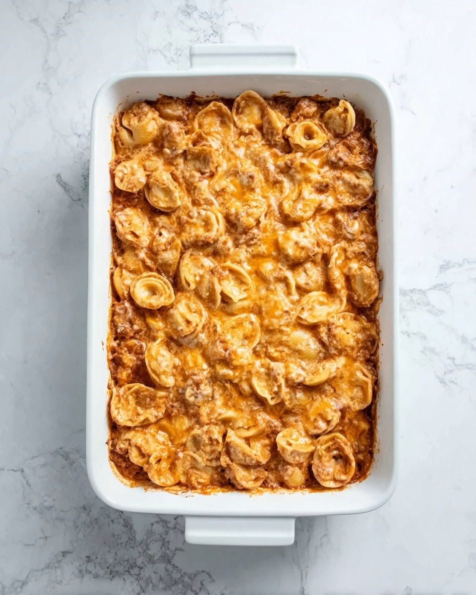 The image shows a white rectangular dish filled with a layered casserole. The top layer is made up of roasted or baked pasta shells that are golden brown with a slightly crispy texture. Below the pasta layer, there seems to be a thick, rich sauce with a smooth texture and a rich orange color, likely cheese or tomato-based. The dish sits on a white marbled surface that adds a clean, bright background. The overall look is warm and hearty, with the pasta shells forming a textured pattern on top, and the sauce layer visible at the edges. photo taken with an iphone --ar 4:5 --v 7