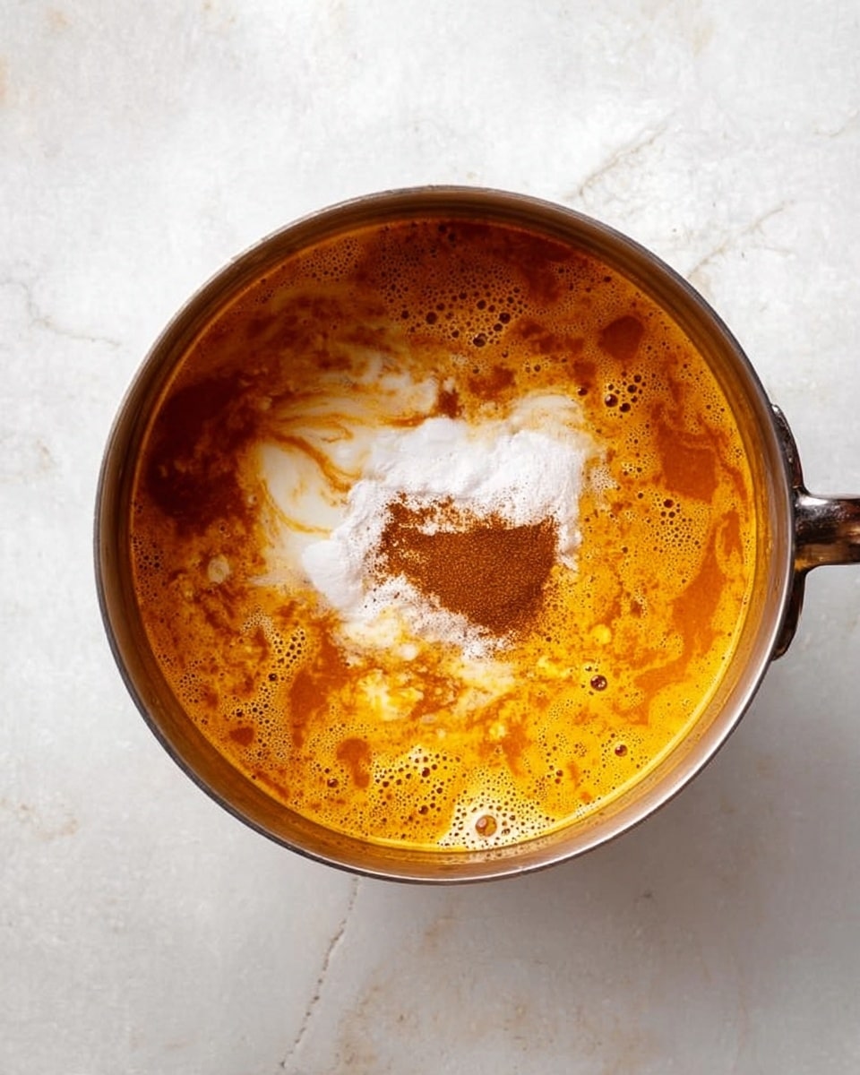 A silver pot sits on a white marbled surface filled with a rich orange liquid that has bubbling areas on the surface. In the center, there is a heap of white powder with a dusting of brown spice on top, blending slightly into the liquid around it. The color shifts from bright orange to deeper brown spots, giving the soup a textured look. Photo taken with an iphone --ar 4:5 --v 7