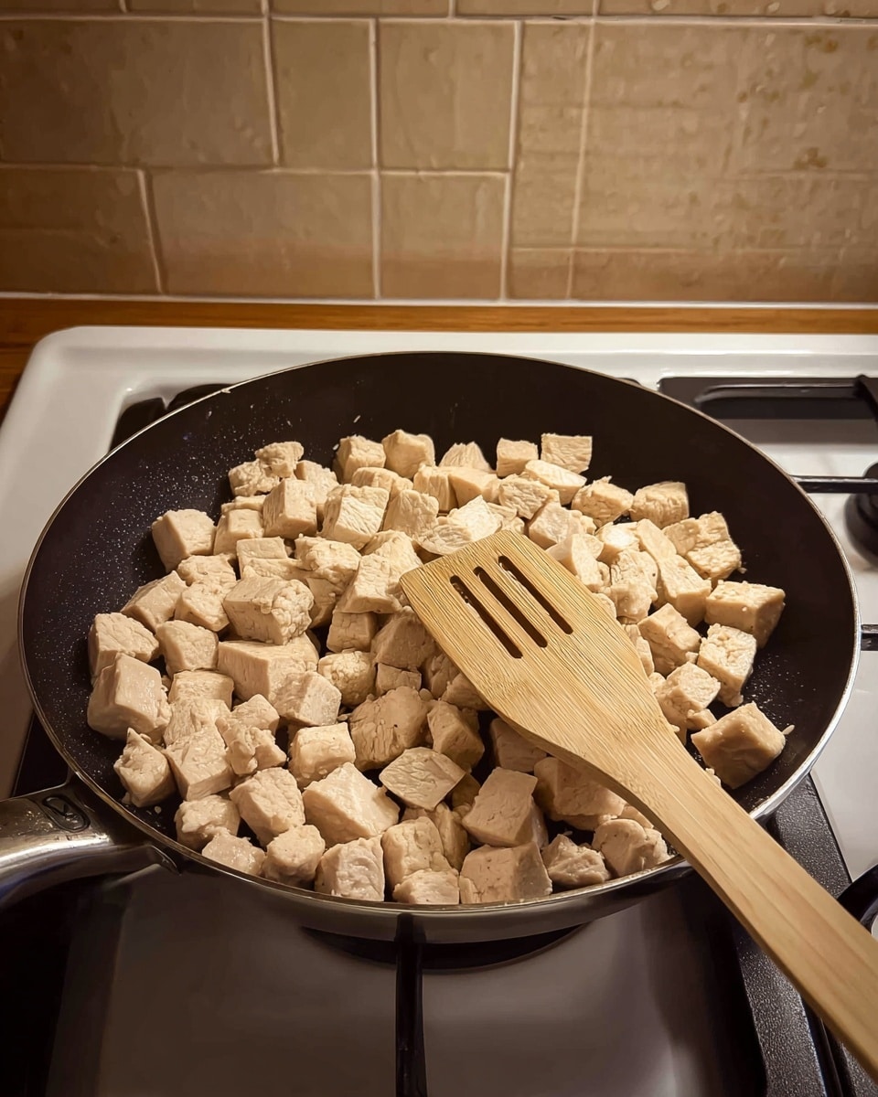 A black frying pan filled with small, pale beige cubes of cooked chicken evenly spread across the surface. A wooden spatula with three long slots rests on top of the chicken cubes, positioned diagonally from bottom right to center. The pan sits on a white stove, and the background shows beige tiled walls and a brown countertop underneath. The scene is well-lit, highlighting the texture of the chicken pieces and the smooth surface of the pan. photo taken with an iphone --ar 4:5 --v 7