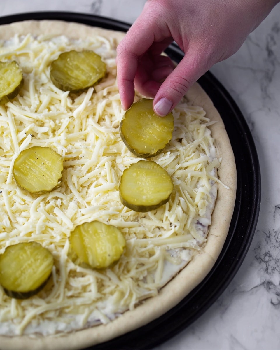A close-up of a round pizza on a black baking tray, placed on a white marbled surface. The pizza has one layer of a creamy white sauce spread evenly over the dough, topped with a thick layer of shredded white cheese. On top of this, several large green pickle slices are being added, with a woman's hand placing one pickle slice carefully. The pizza dough edge is slightly raised and light beige in color. Photo taken with an iphone --ar 4:5 --v 7