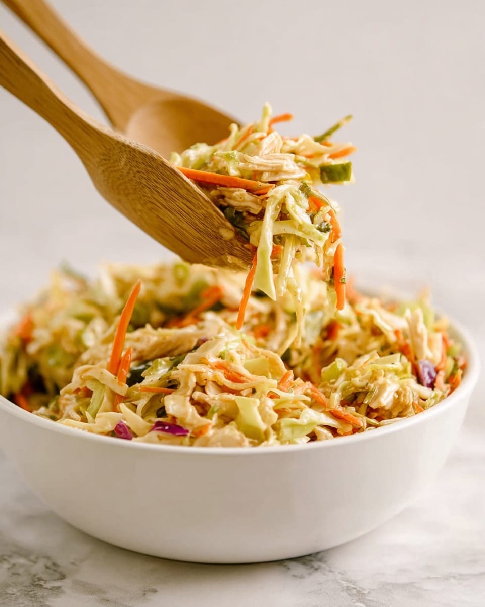 A white bowl filled with a colorful mixed salad is shown with wooden salad spoons lifting a portion above it. The salad has many layers including shredded light green cabbage, thin orange carrot strips, pale beige pieces that look like chicken or tofu, and small pieces of green vegetables scattered throughout. There are also hints of red and purple vegetable bits mixed in, creating a fresh, crunchy texture. The background and surface are a soft white marbled texture, making the colors of the salad pop. The photo is taken with an iphone --ar 4:5 --v 7