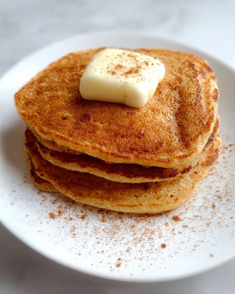 A small stack of four golden brown pancakes sits in the center of a white square plate, each pancake thick and fluffy with smooth edges. One pancake is cut into strips and fanned out slightly to reveal a soft, tender inside with a warm beige color. A silver fork rests on the plate with a few pancake slices on its tines. Light brown cinnamon powder is sprinkled lightly on the plate around the pancakes, adding a touch of texture and color. The background is a white marbled surface. photo taken with an iphone --ar 4:5 --v 7