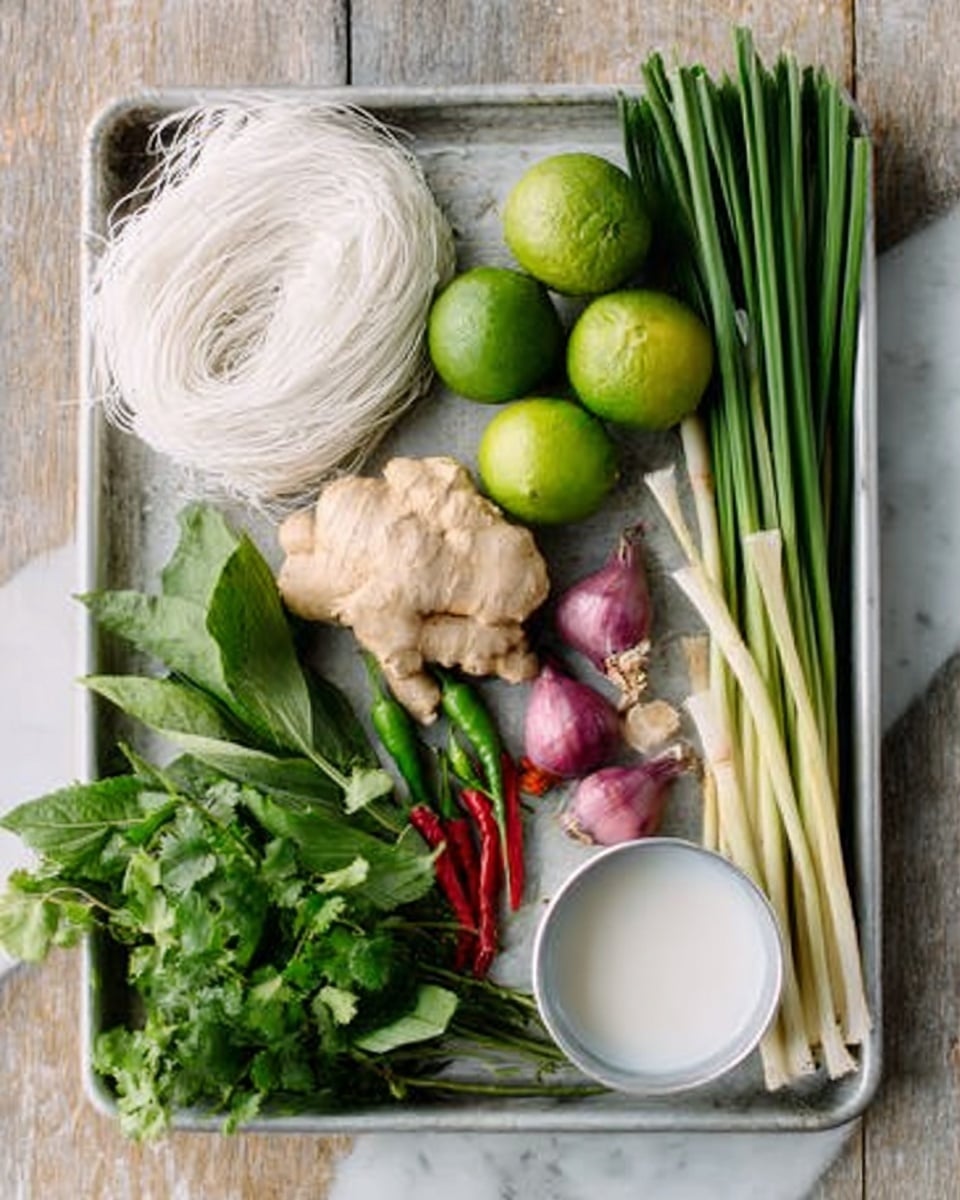 The image shows a white metal tray on a white marbled surface filled with fresh ingredients. There are four light green limes scattered around the tray, along with a large piece of light brown ginger and three small purple shallots grouped near the center. A bundle of thin, white rice noodles is placed on the left side, with a long, pale green lemongrass stalk across the tray diagonally. On the right side, there are several green onions with dark green tops and a few long red and green chili peppers. Fresh green herbs like cilantro and basil fill the bottom left corner, and a small white cup containing a coconut milk-like liquid sits near the bottom right corner. photo taken with an iphone --ar 4:5 --v 7