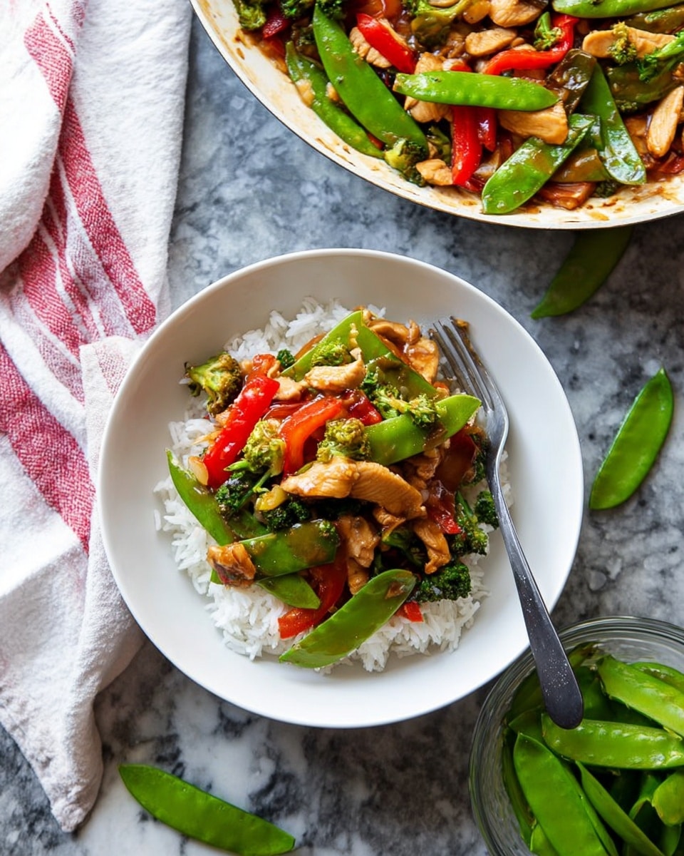 A white pan filled with a stir-fry dish containing several layers: the base layer is a light brown sauce coating the bottom and sides of the pan; on top are small pieces of light brown chicken mixed with bright green snap peas, dark green broccoli florets, and thin slices of red bell pepper; a wooden spoon with sauce and food rests inside the pan. Around the pan are scattered snap peas and a glass container holding fresh snap peas. Two white bowls of cooked white rice are visible, placed on a white marbled surface. A red and white striped cloth is partially visible on the right side. Photo taken with an iphone --ar 4:5 --v 7