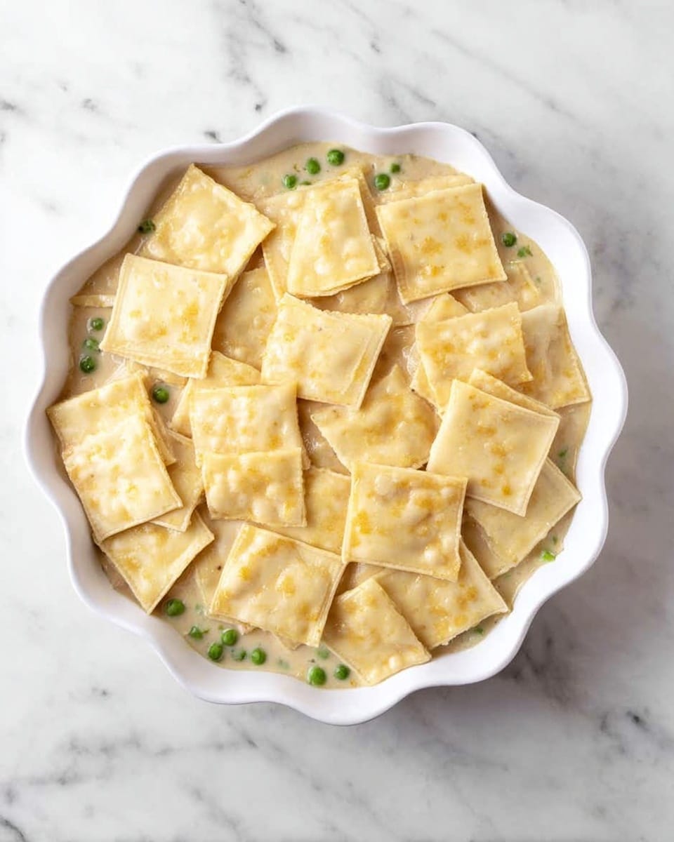 A white scalloped dish with one layer of pale yellow square dough pieces slightly overlapping, placed over a creamy, light tan sauce mixed with small green peas visible underneath. The dough shows slight yellow spots and a soft texture, covering the sauce evenly in the dish. The dish is on a white marbled surface photo taken with an iphone --ar 4:5 --v 7