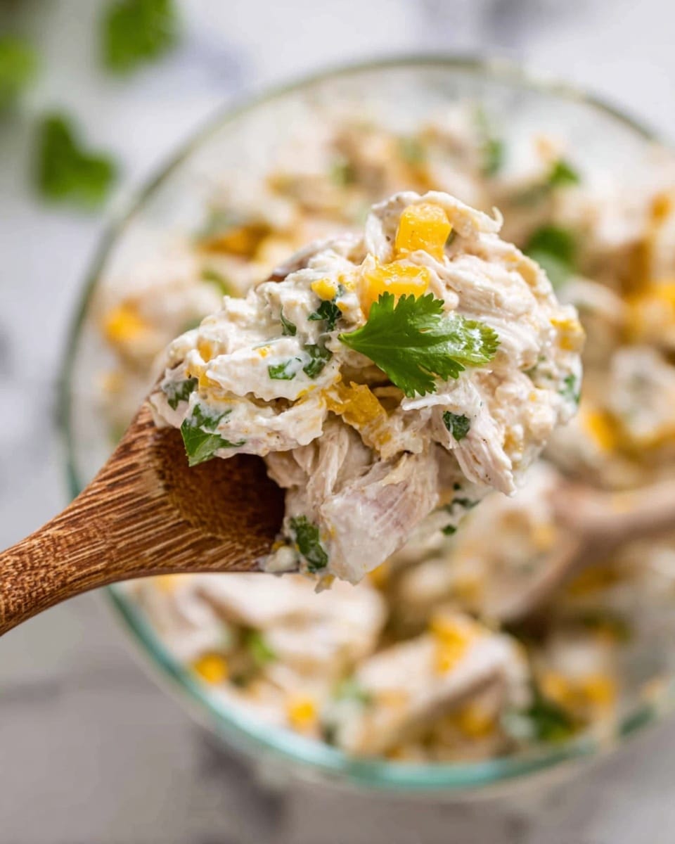 A close-up view of a wooden spoon holding a creamy salad mixture with visible layers of shredded white chicken, small yellow cheese cubes, and fresh green cilantro leaves on top. The background shows a clear glass bowl filled with the same salad, resting on a white marbled surface, with more bits of chicken, cheese, and cilantro mixed together. The texture looks soft and creamy, with the yellow cheese adding bright color contrast against the white chicken and cream. Photo taken with an iphone --ar 4:5 --v 7