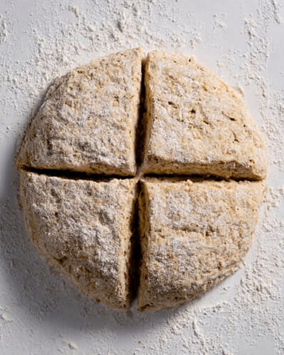 The image shows a round scone dough on a white marbled surface, dusted lightly with white flour. The dough is divided into four equal pieces by two deep cuts crossing in the middle, forming a plus sign. The dough itself has a rough, slightly grainy texture and a light brown color with a matte finish. The surface is clean except for some scattered flour around the dough. Photo taken with an iphone --ar 4:5 --v 7