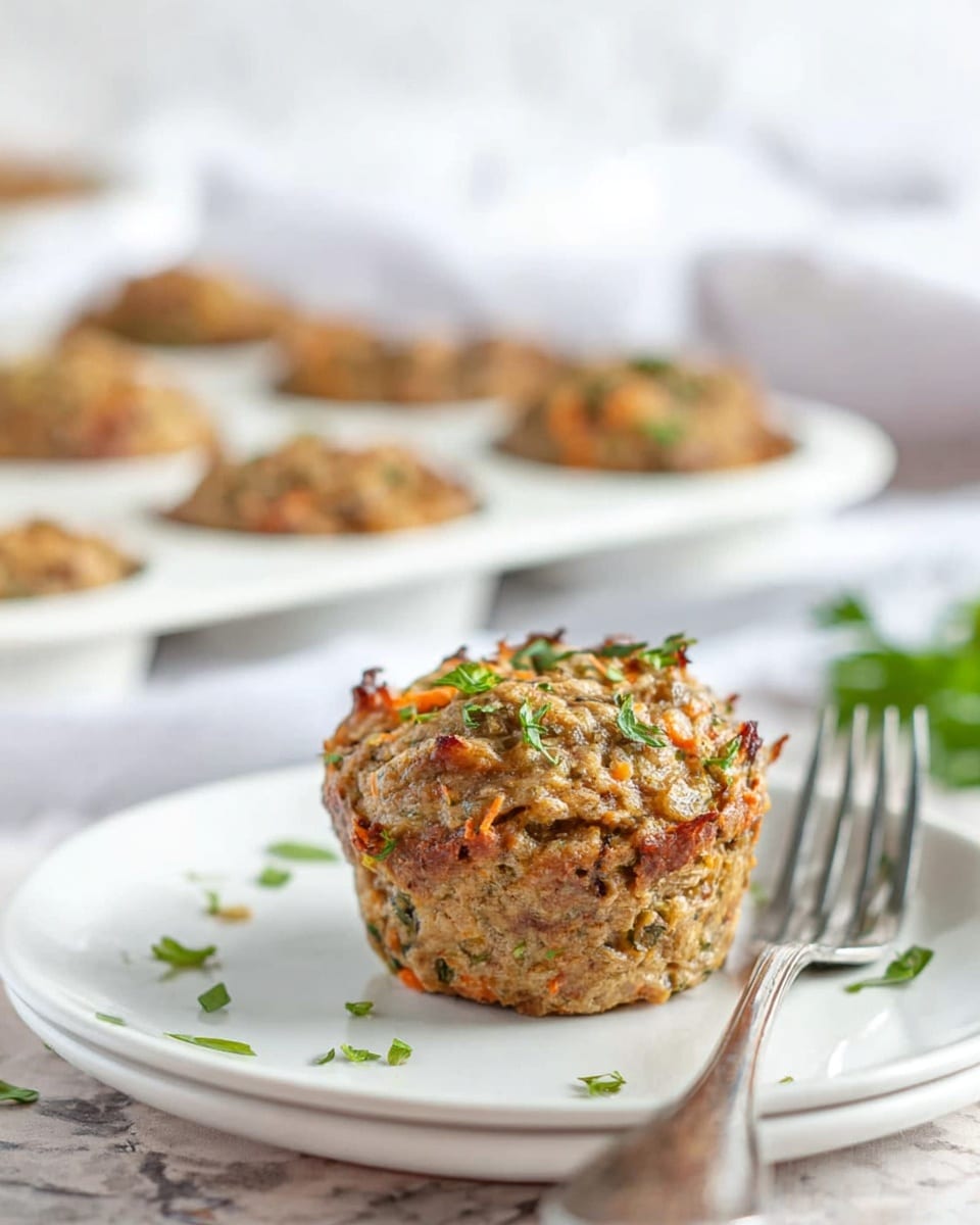 The image shows a single savory muffin on a white plate with a white marbled surface underneath. The muffin has a rough texture with visible small pieces of orange and green herbs mixed inside and sprinkled on top, giving it a colorful, rustic look. Behind the plate, a white muffin tray holds several other muffins with similar appearance that are slightly blurred. There is a silver fork placed on the right side of the plate, and some small green herb pieces scattered around the plate. The background is bright and softly out of focus, highlighting the muffin in the front. Photo taken with an iphone --ar 4:5 --v 7