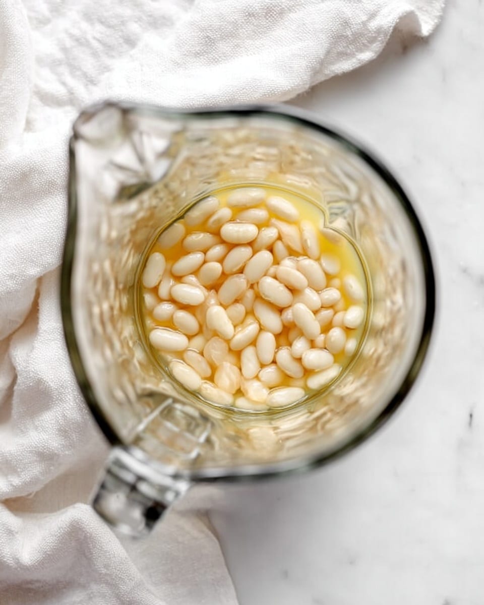 Inside a clear glass blender, small white beans fill about the lower half of the pitcher. The beans sit in a thin layer of light yellow liquid, which covers them partially. The glass blender is placed on a white marbled surface with a soft, white cloth nearby, creating a clean and bright setting. photo taken with an iphone --ar 4:5 --v 7