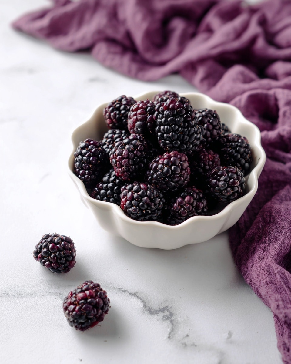 A small white bowl with wavy edges is filled with many dark purple blackberries, each showing a bumpy texture from the tiny drupelets. Around the bowl, three blackberries lie scattered on a white marbled surface. In the background, a soft, crumpled deep purple cloth adds contrast behind the bowl. The light softly shines from the top left, creating gentle shadows and highlights on the berries and bowl photo taken with an iphone --ar 4:5 --v 7