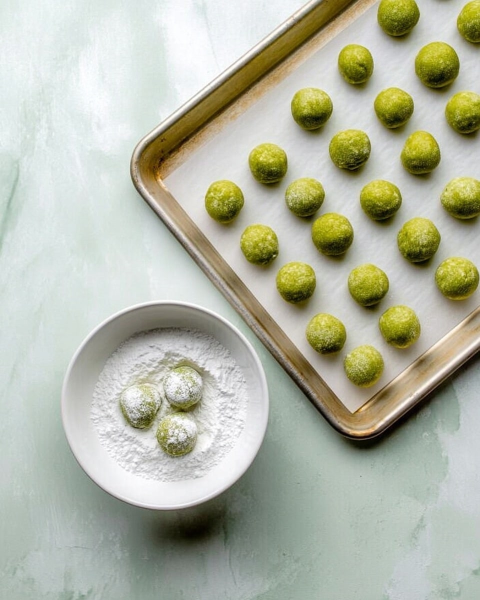 The image shows a silver baking tray lined with white parchment paper filled with many small round green dough balls, evenly spaced. Below the tray, there is a white bowl filled with white powdered sugar, containing three green dough balls partially dusted in the sugar. The background surface is a white marbled texture. A woman's hand is lightly touching one of the dough balls in the bowl. photo taken with an iphone --ar 4:5 --v 7