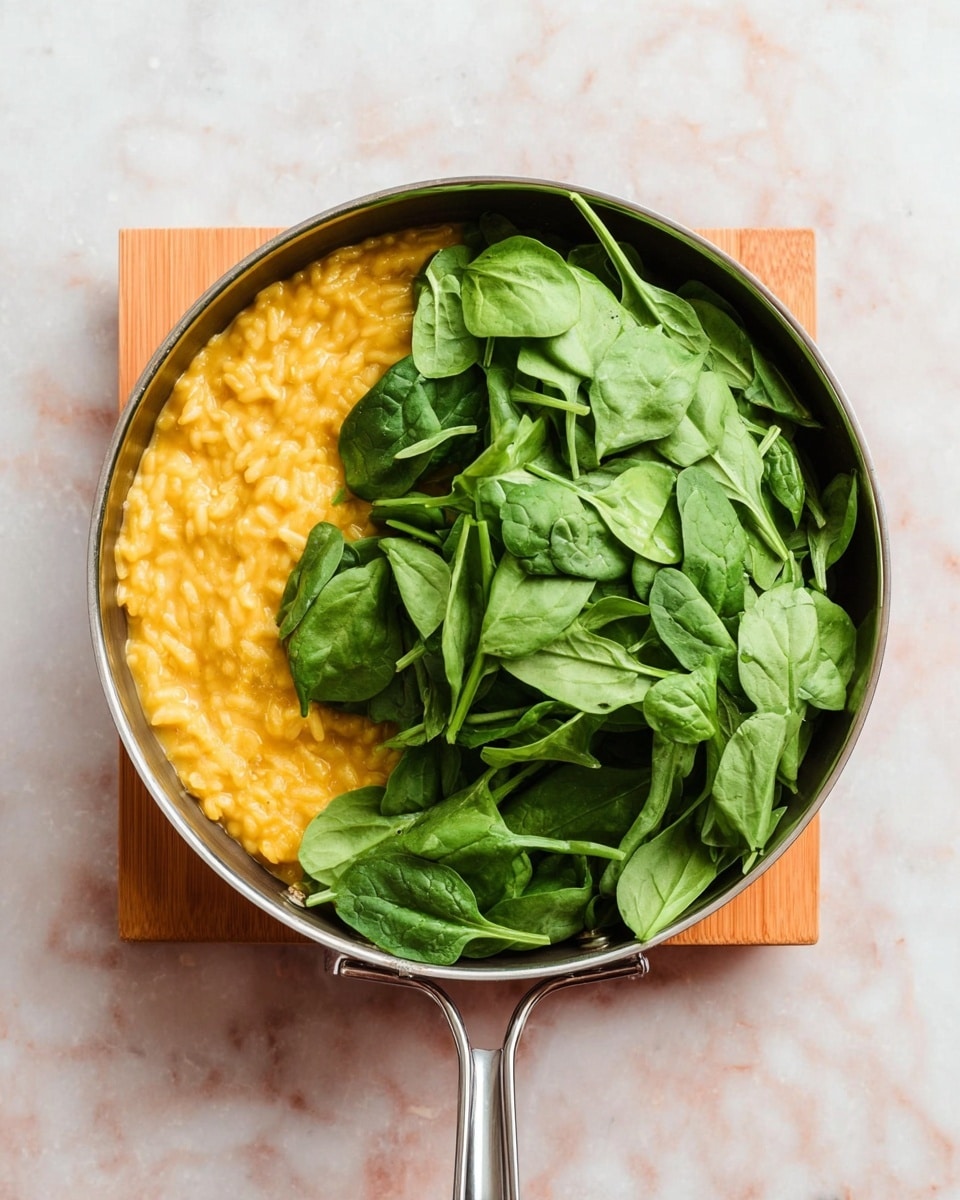 A metal pan placed on a small wooden stand holds two main layers: on the left side, there is a thick, creamy yellow-orange risotto with a smooth texture, and the right side is covered with a large pile of fresh green spinach leaves with visible veins and a slightly glossy surface, sitting loosely in the pan. The pan handle extends downward out of the frame, and the background is a white marbled texture. photo taken with an iphone --ar 4:5 --v 7