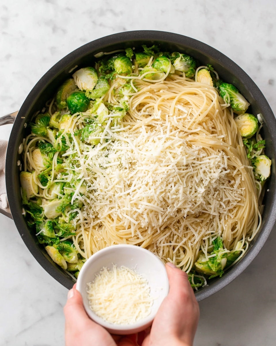 A black pan filled with three layers: the bottom layer is pale yellow spaghetti noodles that are long and thin, spread evenly across the pan; the middle layer has bright green Brussels sprouts cut in half with a slightly cooked texture mixed thoroughly with the noodles; the top layer is soft shredded white cheese being sprinkled from a small white bowl held by a woman's hand over the center of the pan. The background is a white marbled texture. Photo taken with an iphone --ar 4:5 --v 7