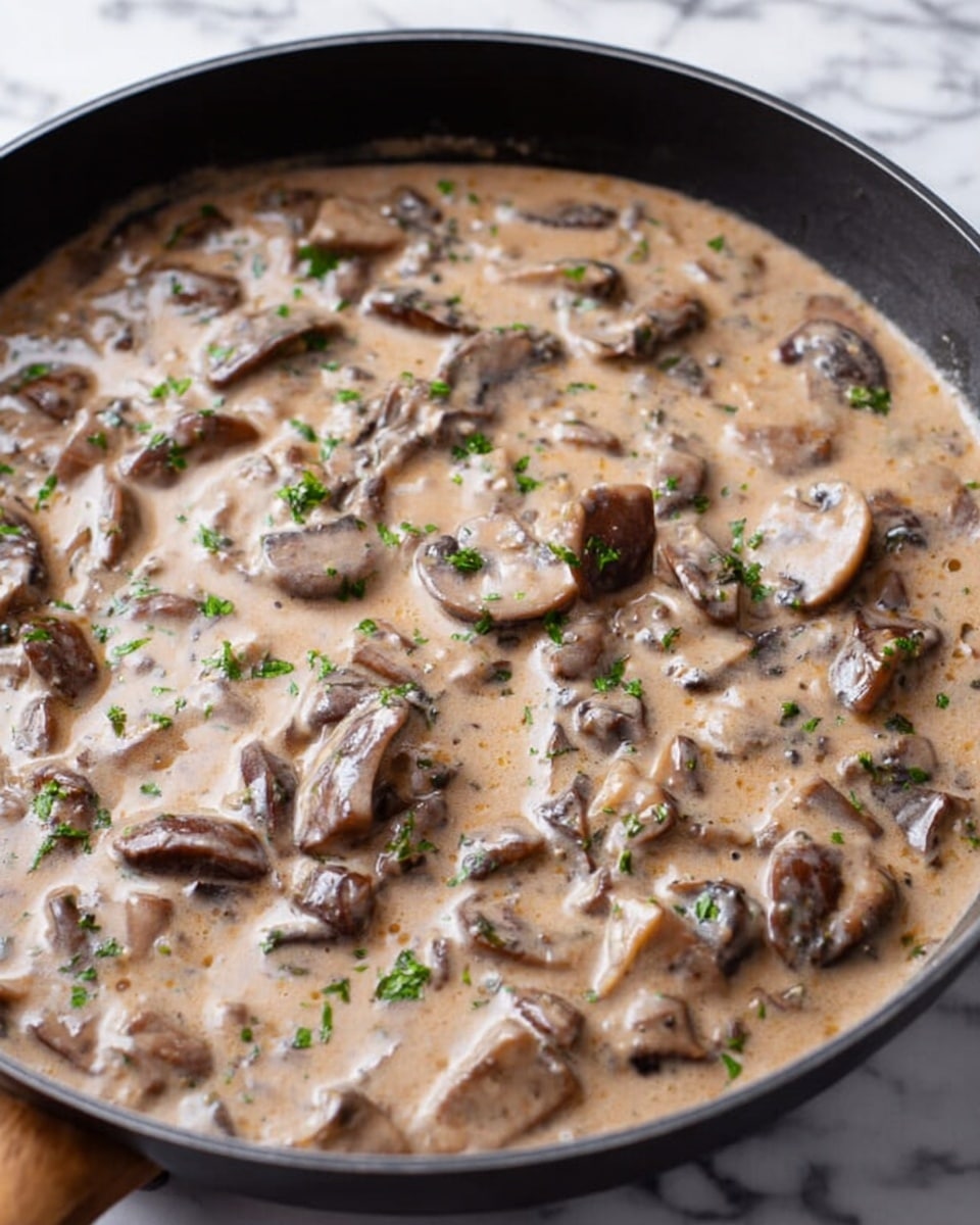 The image shows a black pan filled with a creamy mushroom sauce. The sauce is light brown and thick, with slices of dark brown mushrooms spread evenly throughout. Small pieces of green herbs are sprinkled on top, adding a touch of color and freshness. The pan sits on a white marbled surface, and the sauce looks smooth with some visible texture from the mushrooms. photo taken with an iphone --ar 4:5 --v 7