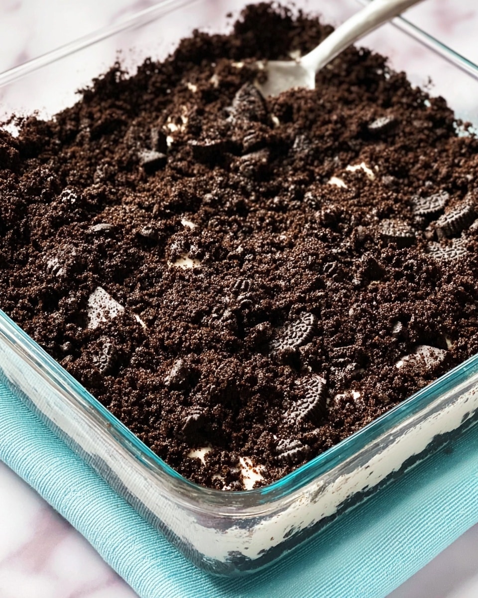 A glass square dish filled with a thick layer of crushed dark chocolate cookies, creating a rough and crumbly texture on top. There are bits of white cream visible inside some cookie pieces, giving small light spots across the dark surface. The dish sits on a light blue cloth, all placed on a white marbled texture. Photo taken with an iphone --ar 4:5 --v 7
