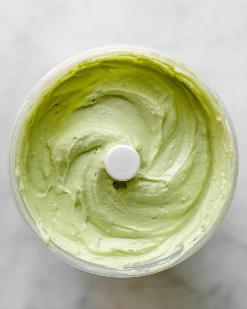 A close-up top view of a round transparent container filled with a smooth light green creamy mixture that has a soft and thick texture, with a white plastic blade in the middle. The container is placed on a white marbled surface. The creamy layer fills the container to about three-quarters of its height, and the mixture is evenly spread, showing slight swirls and ridges on the surface. photo taken with an iphone --ar 4:5 --v 7