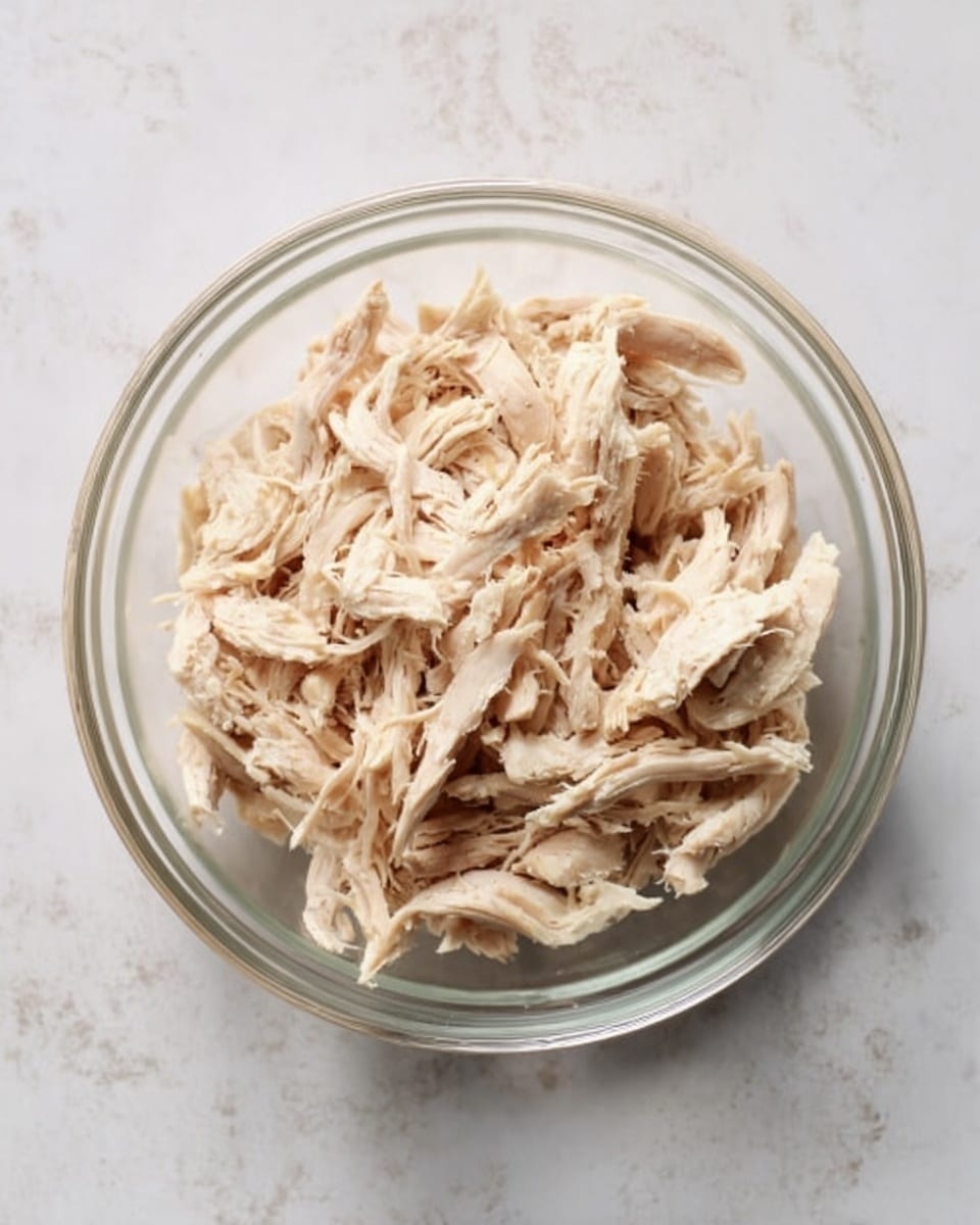 A clear glass bowl filled with shredded cooked chicken. The chicken is light beige and white, with soft and slightly fibrous texture visible. The bowl is placed on a white marbled surface. The lighting is natural and evenly bright. photo taken with an iphone --ar 4:5 --v 7