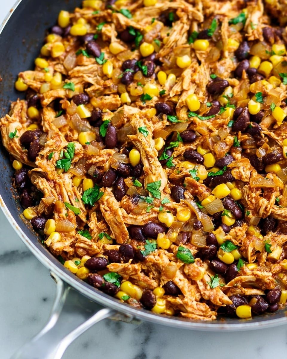 A close-up view of a pan filled with bright yellow corn kernels, dark black beans, and shredded light brown chicken mixed with small pieces of sautéed onions and finely chopped green herbs scattered on top. The textures vary from soft chicken strands to smooth corn and beans, all coated with a light reddish-brown seasoning. The pan sits on a white marbled surface, with a silver handle visible at the edge. Photo taken with an iphone --ar 4:5 --v 7