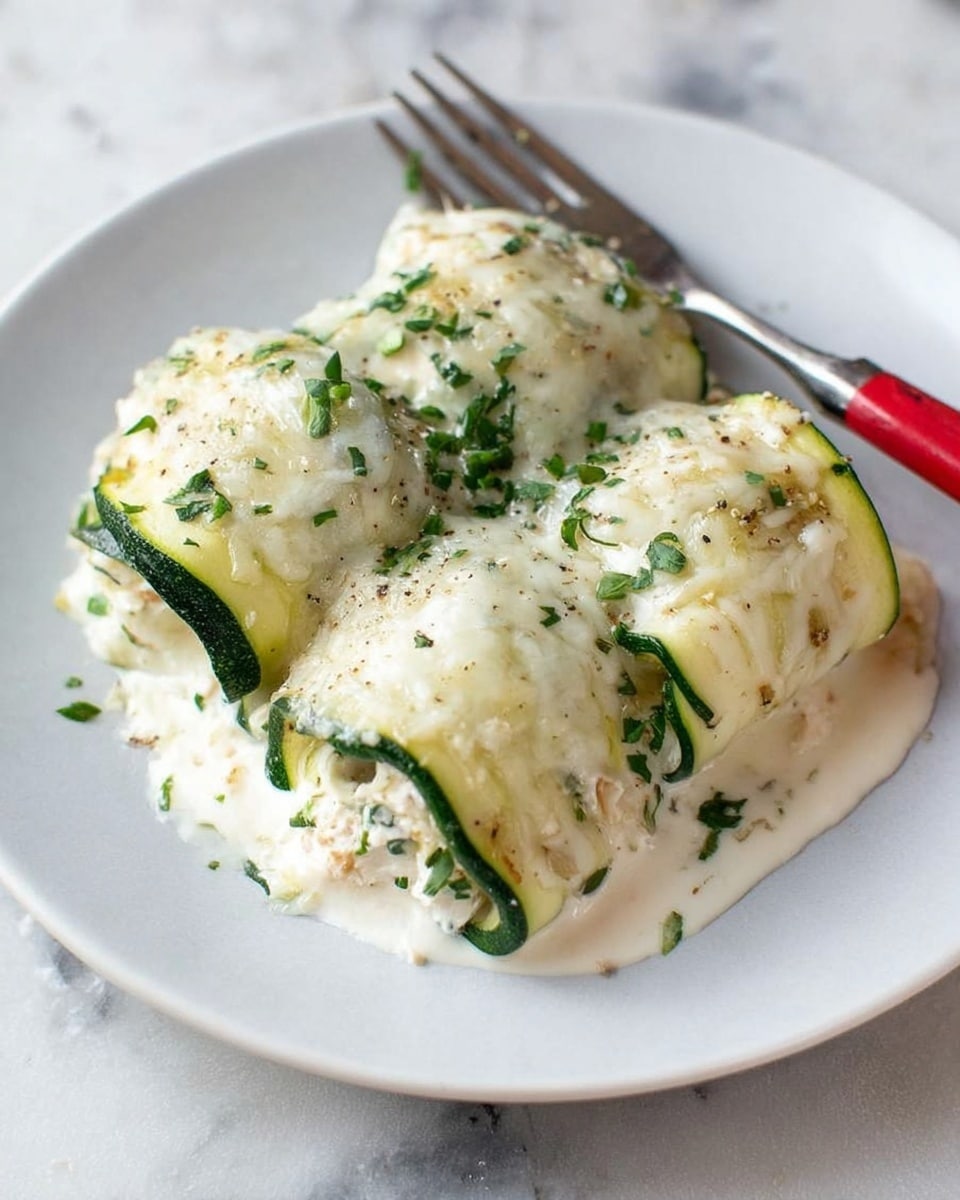 The image shows three rolls made with green zucchini strips wrapped around a creamy white filling, placed on a white plate. Each roll is topped with melted white cheese sprinkled with chopped green herbs, adding texture and color. The creamy white sauce spreads underneath the rolls, blending with the cheese on top. A fork with a red handle is partially inserted under one roll, resting on the plate. The plate sits on a white marbled surface. photo taken with an iphone --ar 4:5 --v 7