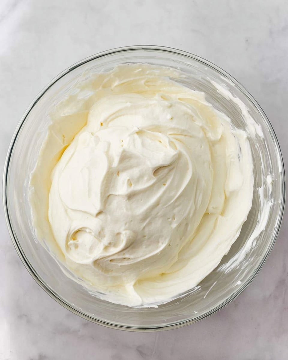 A clear glass bowl filled with a smooth, creamy white mixture that has soft swirls and gentle peaks on the surface. The texture looks light and airy, with some tiny air bubbles and fine specks lightly scattered throughout. The bowl is placed on a white marbled surface. Photo taken with an iphone --ar 4:5 --v 7