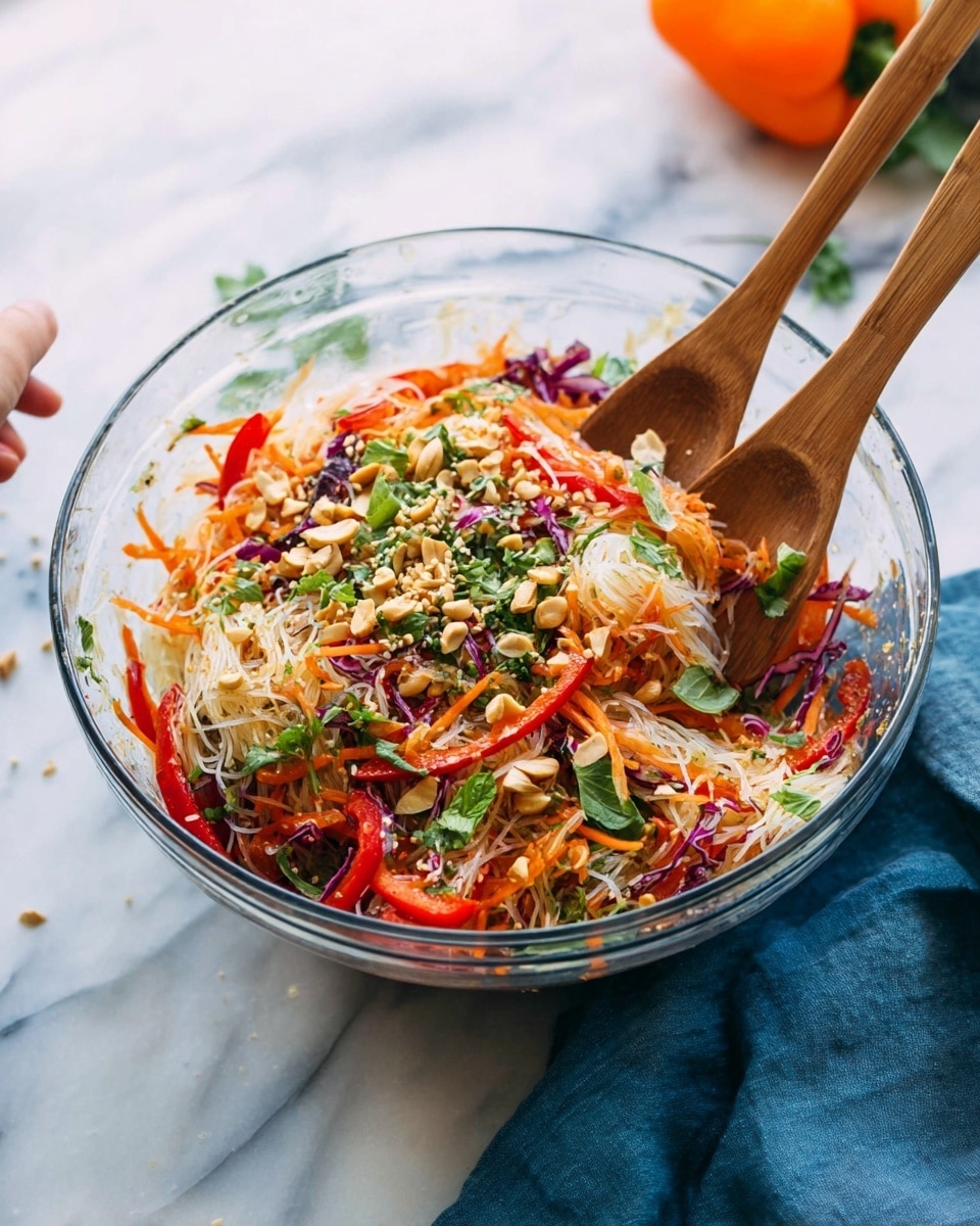 A clear glass bowl holds a colorful layered salad with translucent thin rice noodles at the bottom, topped with julienned bright orange carrots, thin red bell pepper strips, and thin purple cabbage slices. Fresh green herbs and chopped peanuts are sprinkled over the top, adding texture and color contrast. Two wooden spoons rest inside the bowl, one of them touched by a woman's hand. The bowl sits on a white marbled surface with a blue cloth napkin beside it and an orange bell pepper in the background. Photo taken with an iphone --ar 4:5 --v 7