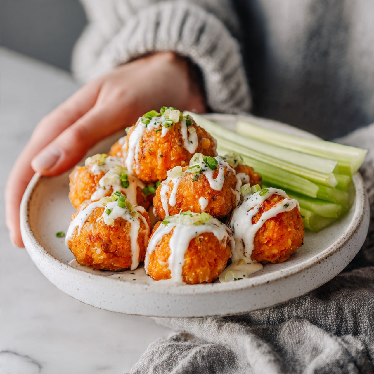 A white round plate on a white marbled surface holds about 15 bright orange, round, crispy balls covered with a drizzle of white creamy sauce. The balls have small green pieces sprinkled on top. On one side of the plate, there are about five long light green celery sticks neatly placed. The plate is partly on a soft gray cloth. A woman's hand holds the edge of the plate from the top left corner. photo taken with an iphone --ar 4:5 --v 7