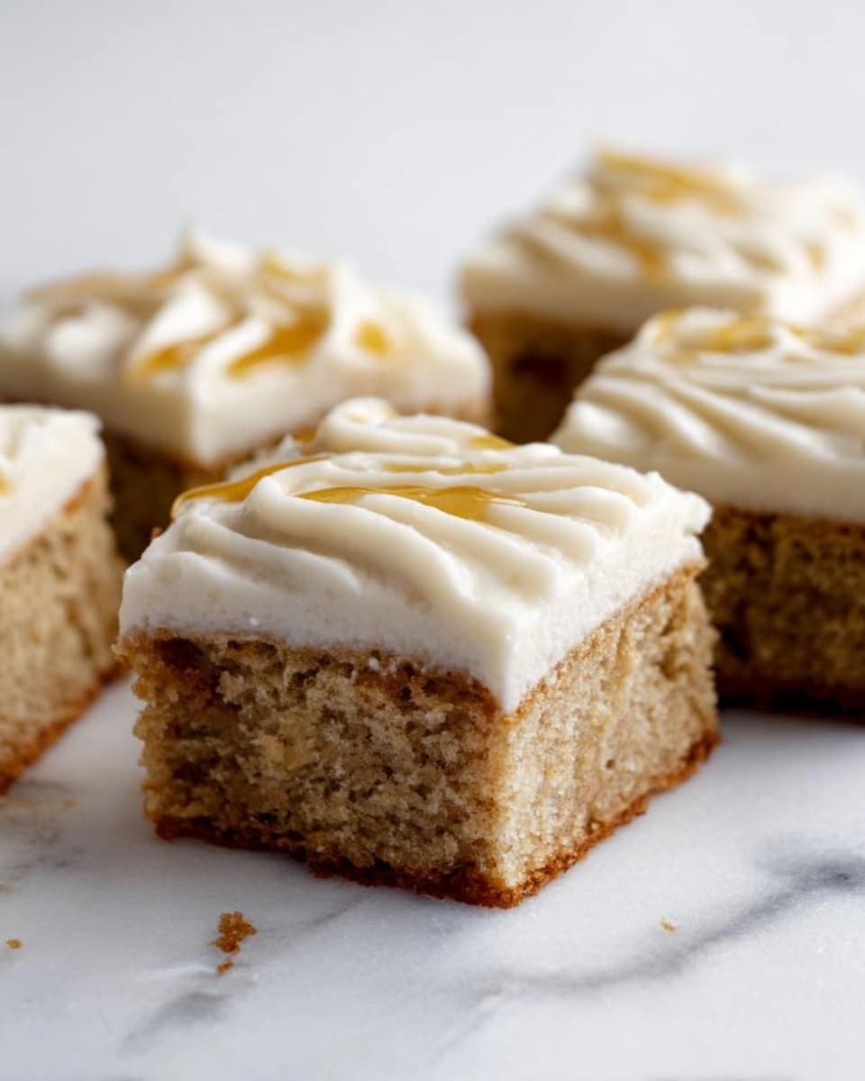 The image shows four square pieces of cake arranged in a row on a white marbled surface. Each piece has two visible layers: the bottom layer is a light brown, moist cake with a slightly rough texture, while the top layer is a thick, creamy white frosting with smooth swirls. A golden syrup drizzle accents the frosting on each piece. The cake looks soft and fresh, with small crumbs scattered around one piece. Photo taken with an iphone --ar 4:5 --v 7