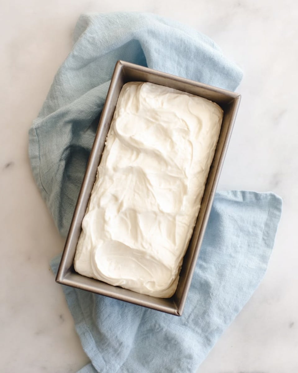 A rectangular white pan filled with creamy white ice cream, unevenly smoothed on top with soft swirls and dips. A metal ice cream scoop rests in the pan, partly buried in the ice cream. Next to the pan is a small white bowl holding two rounded scoops of the same white ice cream. The pan and bowl sit on a white marbled surface. A soft, light blue cloth is loosely placed to the right side of the pan. photo taken with an iphone --ar 4:5 --v 7
