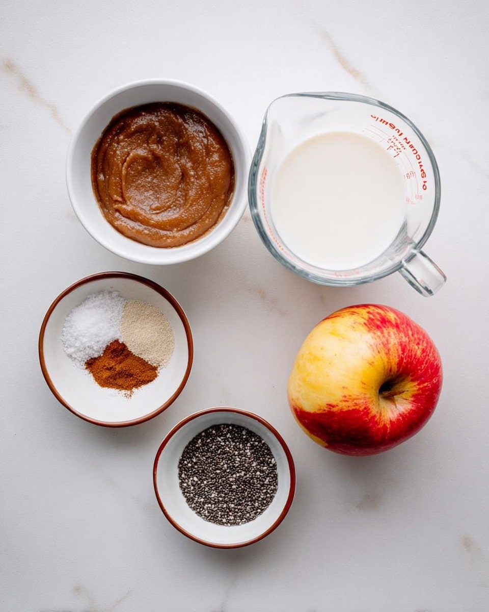 The image shows six ingredients arranged on a white marbled surface. At the top right, there is a clear glass measuring cup filled with white milk. To the left of the milk, there is a white bowl filled with a smooth brown paste, likely apple butter. Below this bowl, there are two small white bowls with brown rims; one contains dark brown and light brown ground spices and white salt, while the other is filled with small black chia seeds. On the far right, next to the milk cup, there is a shiny red and yellow apple. The setup is clean with clear colors and textures of each ingredient. Photo taken with an iphone --ar 4:5 --v 7