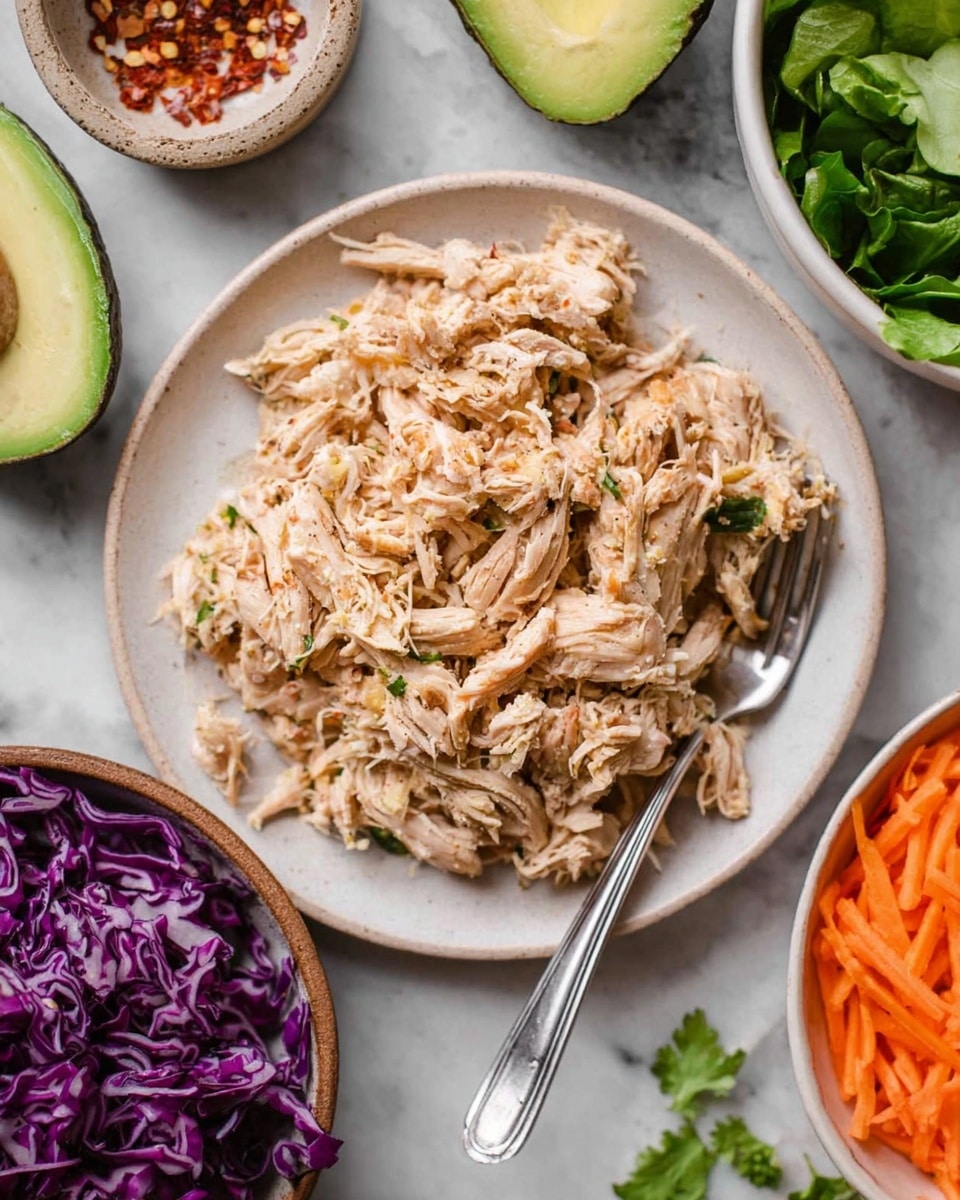 A white plate filled with shredded light brown cooked chicken that looks soft and moist, with a silver fork resting on the right side of the chicken. Around the plate, there is a half avocado with green flesh and a large seed cavity at the top, a bowl of shredded purple cabbage, a bowl of shredded orange carrots, and some fresh green leafy vegetables placed on a white marbled surface. A small bowl with red chili flakes is partially visible in the upper left corner. photo taken with an iphone --ar 4:5 --v 7
