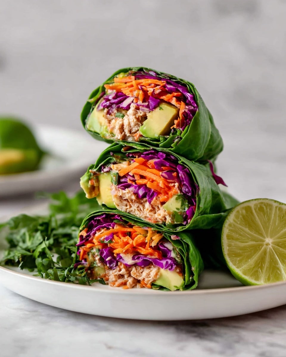Three fresh wraps are stacked on a white plate, each wrapped in bright green leaves. Inside the wraps, there are layers of purple cabbage, orange shredded carrots, chunks of light green avocado, and a beige filling that looks like shredded chicken or tuna mixed with sauce. On the side of the plate, there is a lime half and some green herbs, all placed on a white marbled surface. The photo is taken with an iphone --ar 4:5 --v 7