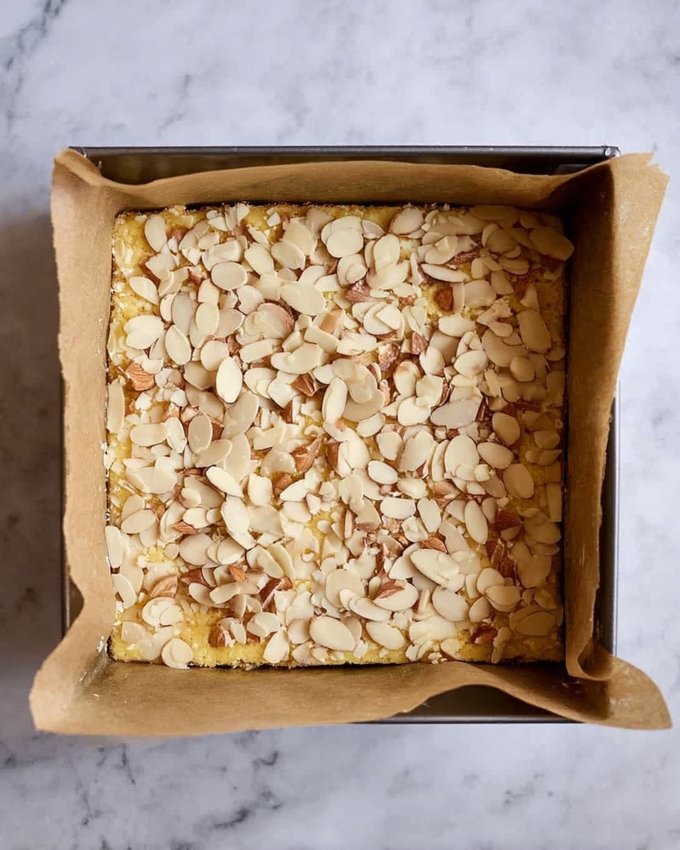 The image shows a square baking pan lined with brown parchment paper on two opposite sides. Inside the pan, there is a single thick yellowish cake layer topped with many sliced almonds spread evenly across the surface, creating a textured and nutty look. The pan sits on a white marbled surface. photo taken with an iphone --ar 4:5 --v 7