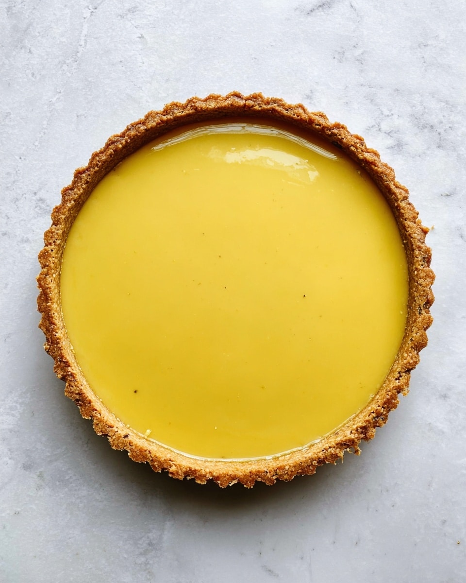 A round tart with one main layer of smooth, shiny yellow filling that covers the entire center. The crust around the tart is light brown, thick, and has a slightly rough texture with small dark spots. The tart is placed on a surface with a white marbled texture. photo taken with an iphone --ar 4:5 --v 7