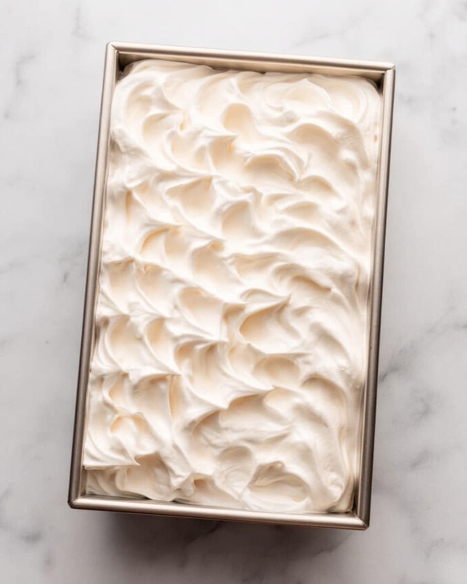 The image shows a rectangular metal pan filled with a layer of smooth, fluffy white meringue spread evenly on top. The meringue has soft, wavy peaks and ridges across the surface, giving it a light and airy texture. The pan is placed on a white marbled surface that contrasts with the pale creamy color of the meringue. photo taken with an iphone --ar 4:5 --v 7