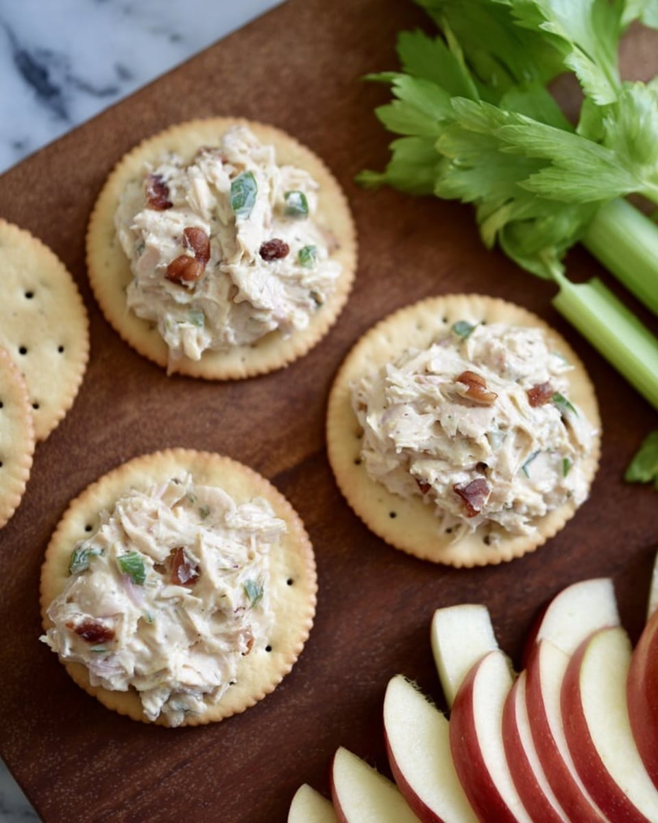 The image shows four round crackers on a wooden board with a white marbled background. Three of the crackers have a thick layer of chicken salad on top, with the salad looking creamy and mixed with small green herb pieces and bits of bacon or nuts. The crackers are a light golden color with small holes on their surface. Toward the top right, fresh celery stalks with green leaves appear, and on the bottom right, there are thinly sliced pieces of red apple with white inner flesh. The scene looks fresh and simple. Photo taken with an iphone --ar 4:5 --v 7