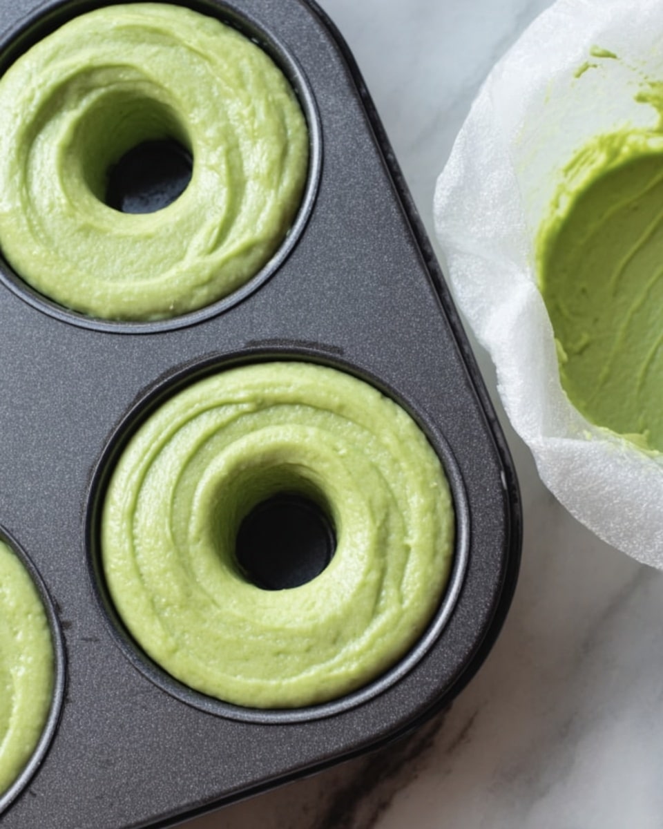 A close-up image showing a dark metal donut baking tray with one donut cavity filled with bright green batter. The batter is smooth and swirled inside the round donut shape, with a clean circle in the middle where the hole will be. Next to the tray is a white icing bag filled with the same green batter, positioned to the right side, ready for more filling. The tray rests on a white marbled surface. photo taken with an iphone --ar 4:5 --v 7