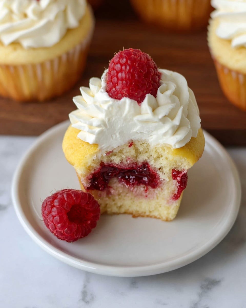 A cupcake is shown from above on a plain white plate set on a white marbled texture. The cupcake has three visible layers: a soft yellow base with a deep red raspberry jam center, a thick layer of whipped white cream on top, and a single fresh red raspberry placed in the center of the cream. Another whole raspberry sits on the plate beside the cupcake. Part of two additional cupcakes topped with whipped cream and a raspberry are visible at the edges of the image. Photo taken with an iphone --ar 4:5 --v 7