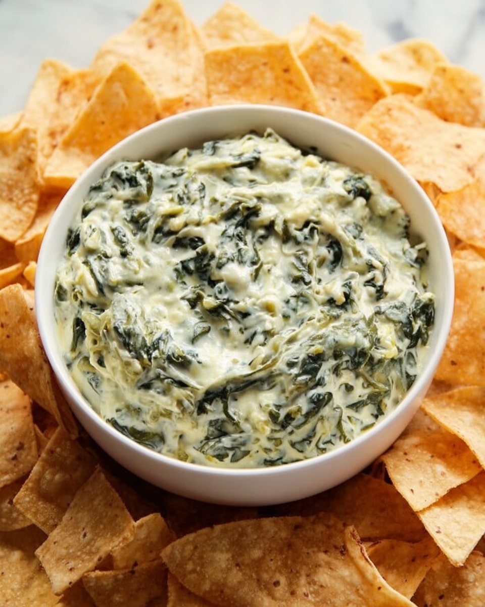 A white bowl filled with a creamy spinach dip that has a thick, textured mix of green spinach leaves and white creamy sauce, sitting in the middle of a circle of light brown tortilla chips. The chips are triangular and look crispy, covering a white marbled surface. The dip appears slightly chunky with visible bits of spinach evenly spread throughout. photo taken with an iphone --ar 4:5 --v 7