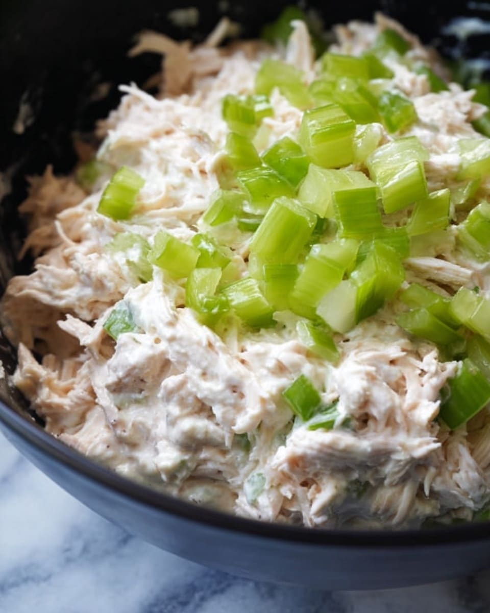 The image shows a close-up of a black bowl filled with a creamy mixture of shredded light pink chicken and white mayo, with bright green chopped celery pieces on top. The texture of the chicken and mayo is smooth and thick, while the celery adds a crunchy contrast. The background is a white marbled texture. photo taken with an iphone --ar 4:5 --v 7