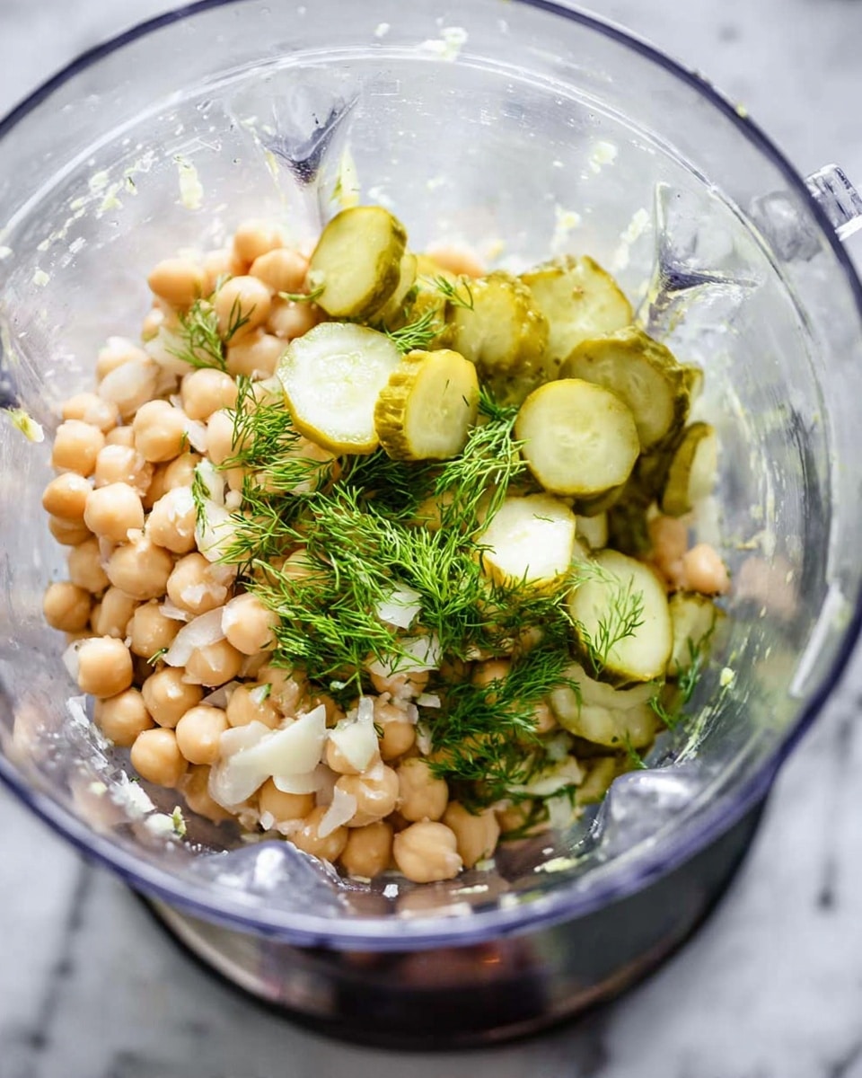 In a clear blender bowl, there is a mixture with three main layers visible: at the bottom, small beige chickpeas form a dense base; on top of this, there are round green pickle slices scattered unevenly, adding a bright contrast; and sprinkled around are small white pieces of garlic and a few fresh, feathery dill sprigs with a bright green color. The blender bowl rests on a shiny metal base, and the overall scene is set against a white marbled texture. photo taken with an iphone --ar 4:5 --v 7