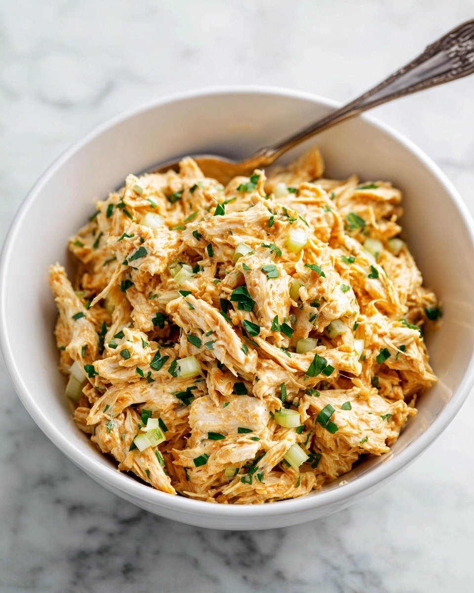 A white bowl filled with a creamy chicken salad that has three main layers visible: shredded light beige chicken mixed with creamy orange sauce, small pieces of pale green celery scattered throughout, and tiny bits of white corn or onion adding texture. Chopped green herbs are sprinkled on top, adding color contrast. A silver spoon rests on the right side inside the bowl, partly covered with the chicken mixture. The bowl sits on a white marbled surface. Photo taken with an iphone --ar 4:5 --v 7