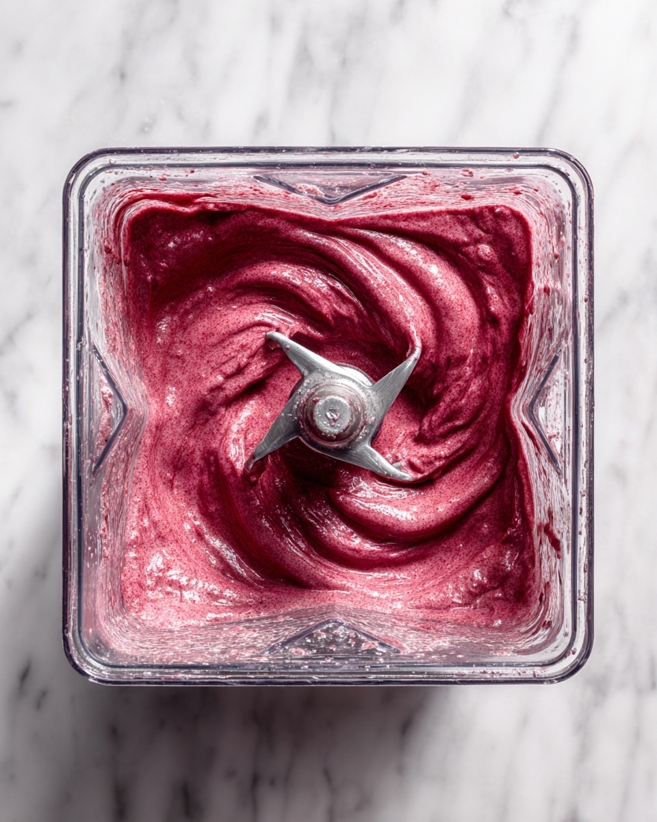 A close-up view inside a blender shows a thick, smooth mixture of deep reddish-purple color swirling around the sharp silver blades at the center. The mixture looks creamy and richly textured, filling the square, clear blender container almost to the top edges. The background is a white marbled surface, giving a clean and bright look to the image. photo taken with an iphone --ar 4:5 --v 7
