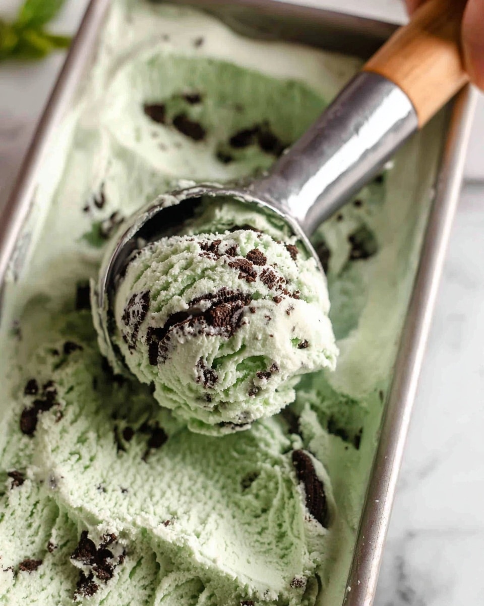 A waffle bowl filled with three scoops of light green mint chip ice cream sprinkled with dark chocolate cookie crumbs, with one whole dark chocolate sandwich cookie stuck on the side of the scoops; a silver spoon is holding a scoop in the background, and a fresh green mint leaf sits beside the bowl on a white marbled surface with scattered cookie pieces around it, photo taken with an iphone --ar 4:5 --v 7