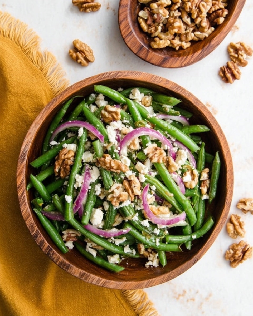 A wooden bowl filled with a green bean salad sits on a white marbled surface. The salad has three layers: the bottom layer is bright green beans cut into short pieces, the middle layer is thin slices of red onion scattered evenly, and the top layer is crumbled white cheese mixed with small pieces of light brown walnuts. Near the bowl, there is a small wooden dish filled with whole walnuts and some walnuts scattered on the white marbled surface. A mustard yellow cloth with frayed edges is partially visible at the bottom left side of the image. Photo taken with an iphone --ar 4:5 --v 7