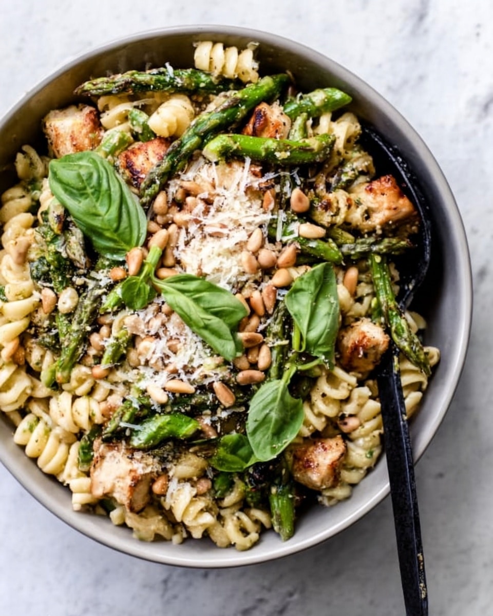 A close-up of a white bowl filled with a pasta dish layered with spiral rotini pasta coated in green pesto sauce, topped with grilled chunks of chicken that have a light brown char, roasted asparagus pieces with a bright green and slightly crisp texture, sprinkled with grated white cheese and scattered golden pine nuts, all garnished with thin strips of fresh green basil leaves, placed on a white marbled surface. Photo taken with an iphone --ar 4:5 --v 7