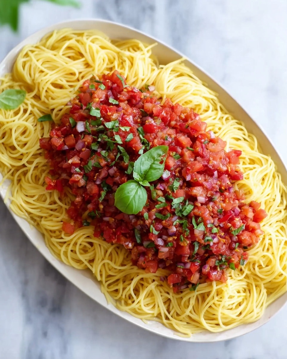 A white oval plate shows a bed of thin yellow spaghetti noodles spread evenly, on top of which is a thick layer of finely chopped tomatoes mixed with small bits of red onion and herbs, forming a chunky red and purple topping that covers the center of the pasta. Fresh green basil leaves are scattered on the tomato mixture, adding pops of green color. The plate is placed on a white marbled surface. photo taken with an iphone --ar 4:5 --v 7