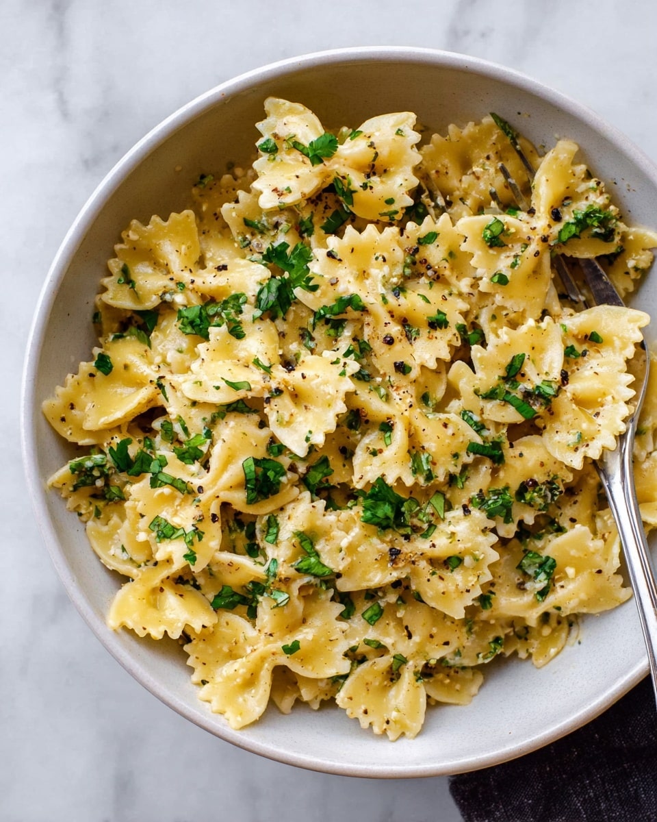 A white bowl filled with bowtie pasta coated in a creamy light yellow sauce. The pasta pieces have ruffled edges and are spread evenly in the bowl. Bright green chopped herbs are sprinkled on top, adding pops of color. Small black pepper flakes are visible on the pasta and herbs. A silver fork rests on the side inside the bowl. The bowl is placed on a white marbled surface photo taken with an iphone --ar 4:5 --v 7