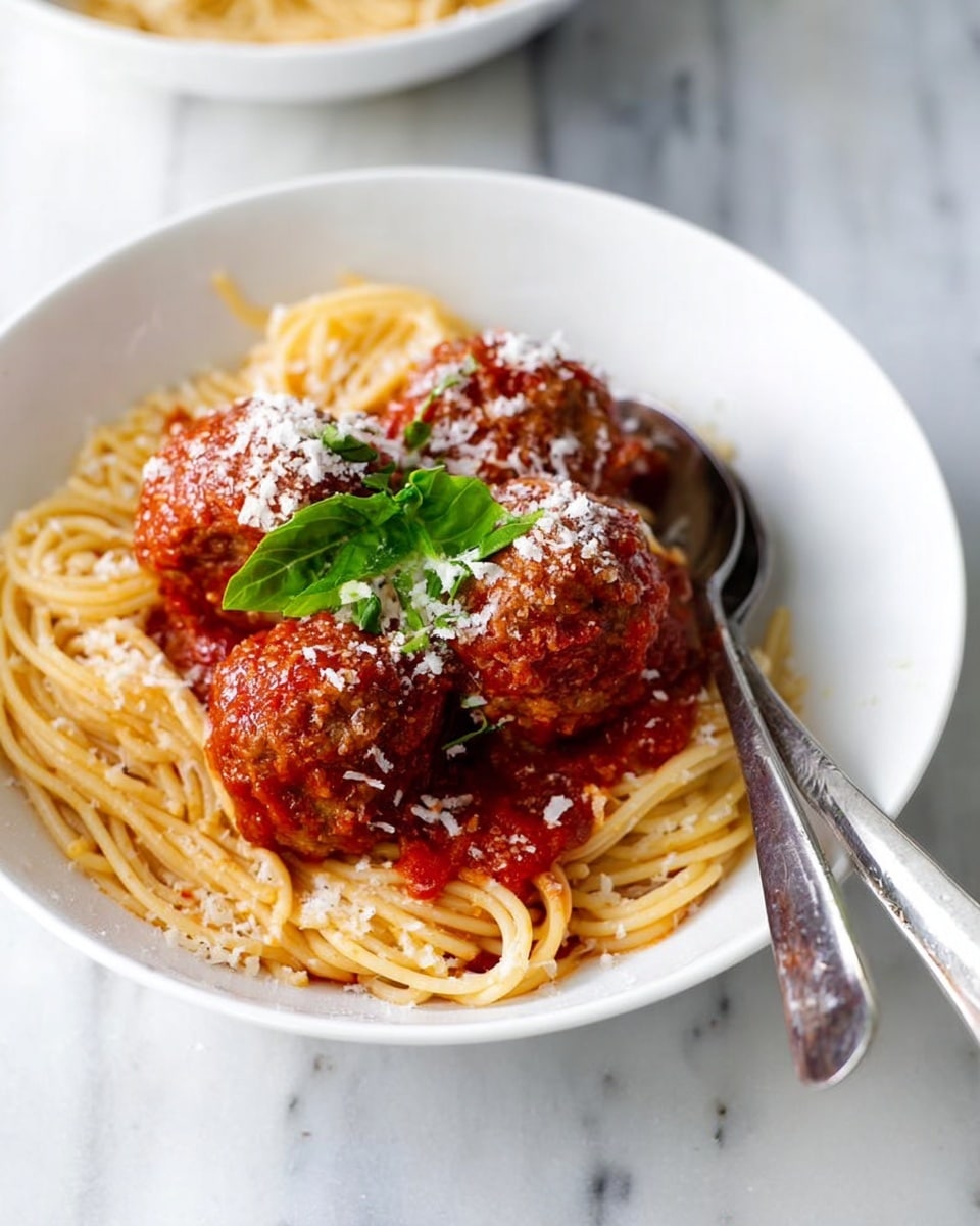 The image shows a white bowl filled with a nest of spaghetti at the bottom, light golden in color with a smooth texture. On top, there are several round meatballs covered in bright red tomato sauce. The meatballs have a slightly rough texture, and the tomato sauce looks thick and rich. The dish is sprinkled with white grated cheese, scattered evenly over the meatballs and sauce. A fresh green basil leaf is placed on top as garnish. A shiny metal fork and spoon rest on the right edge of the bowl. The bowl is set on a white marbled surface. Photo taken with an iphone --ar 4:5 --v 7