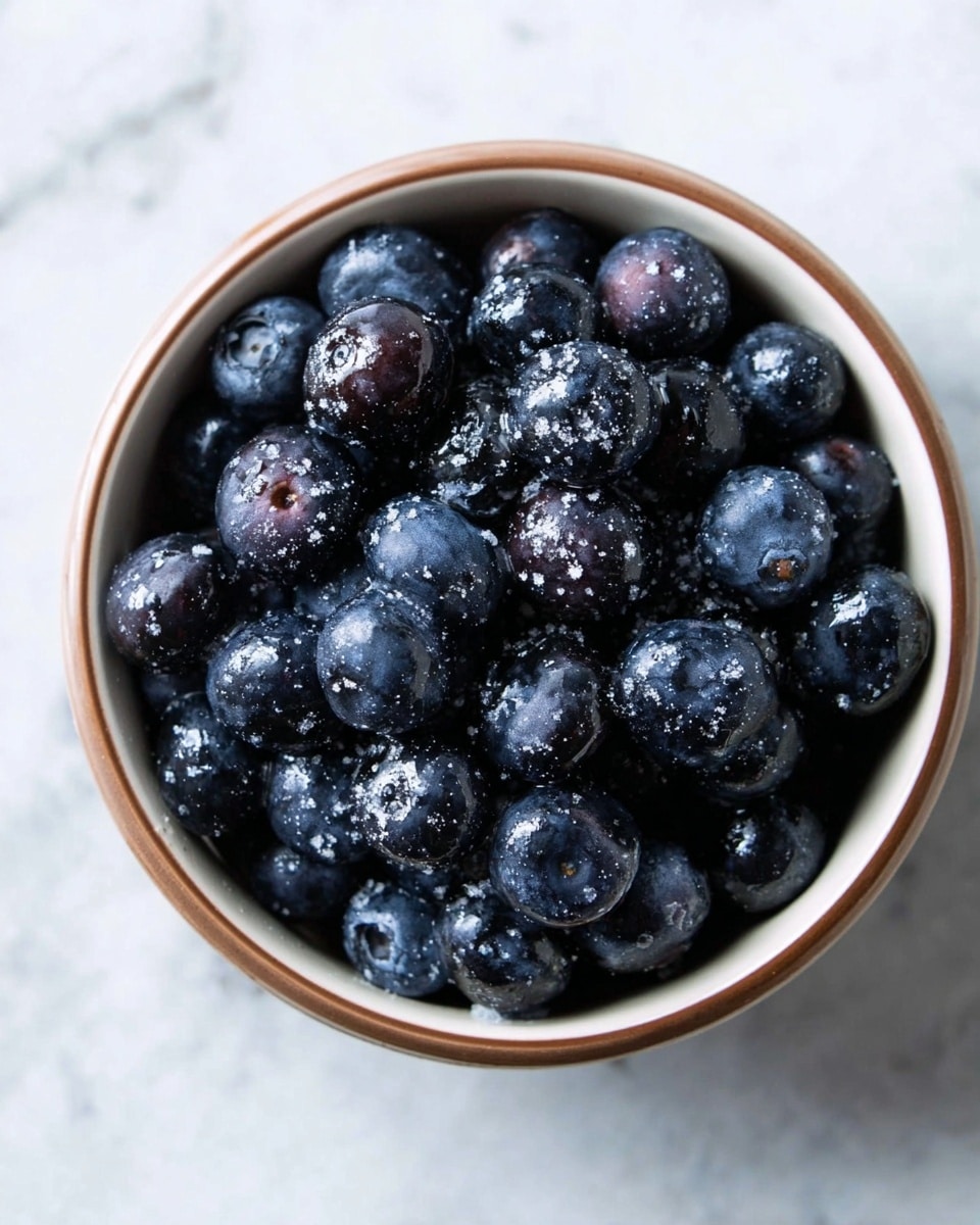 A white bowl filled with dark blue to almost black blueberries that are shiny and fresh looking, some berries sprinkled lightly with white powdered sugar. The bowl has a brown rim and sits on a white marbled surface with soft, natural light highlighting the berries' round shapes and textures. photo taken with an iphone --ar 4:5 --v 7
